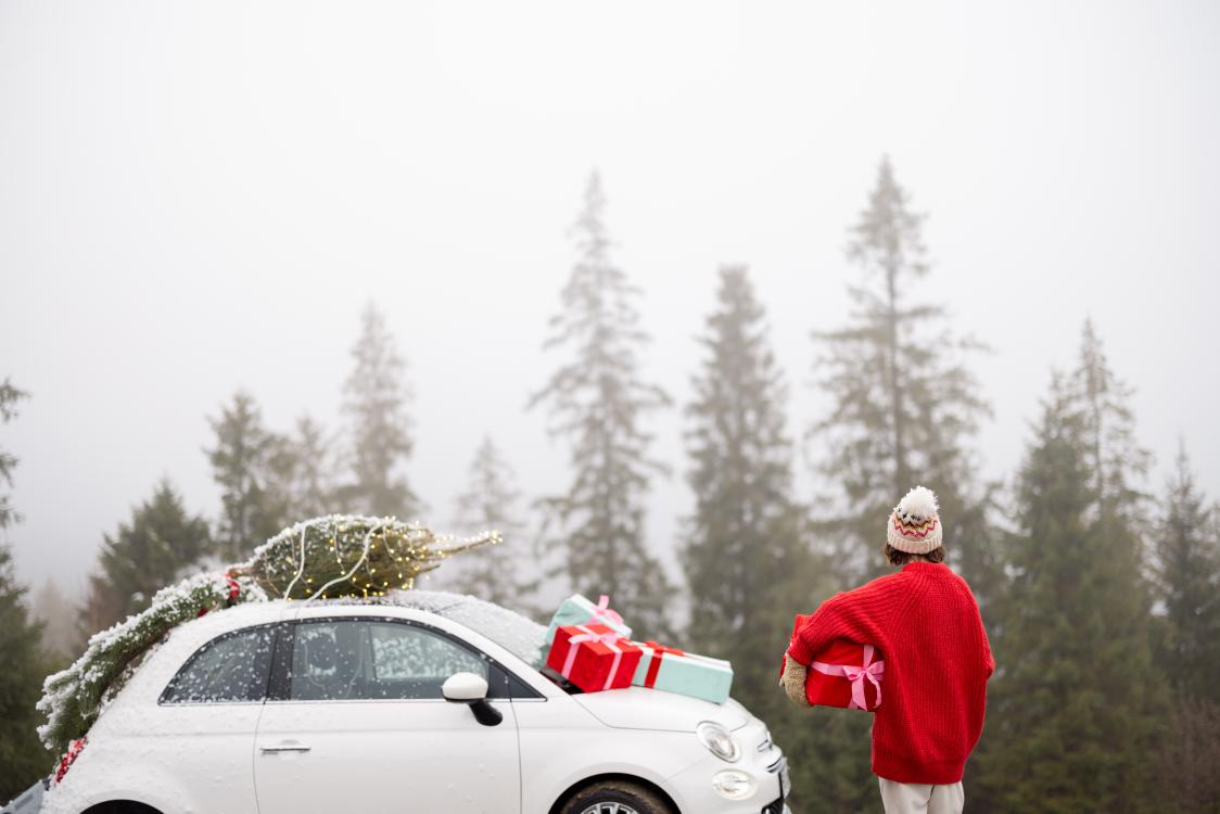 Young woman in red sweater enjoys great view on pine forest, while traveling by car in mountains during winter holidays. Vehicle with Christmas tree, wreath and gift boxes.